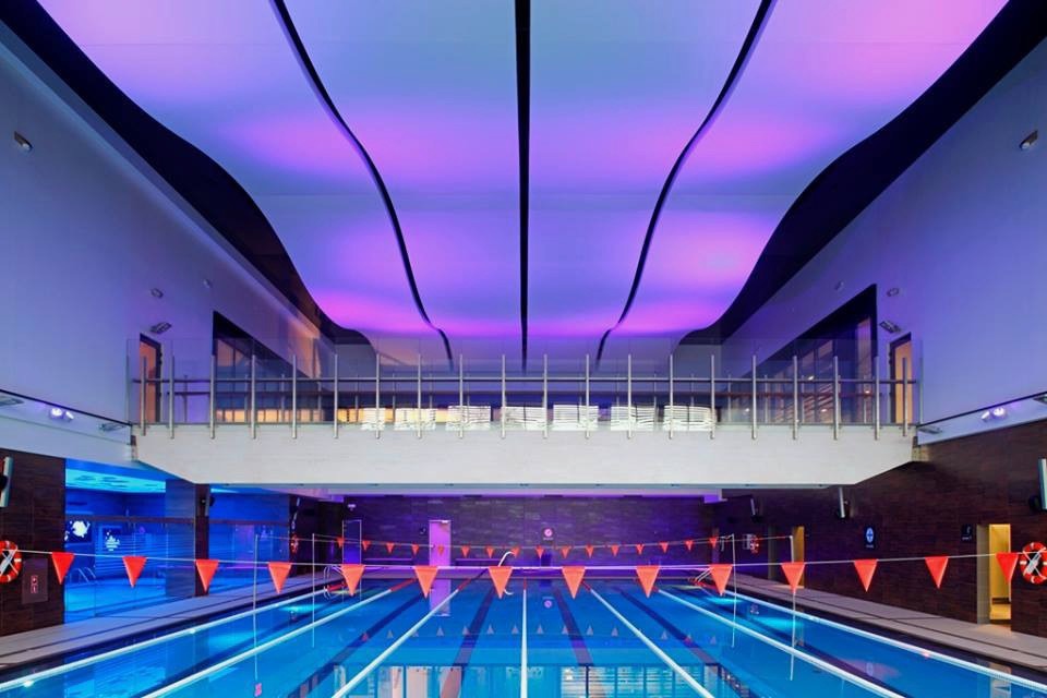 Nowoczesny basen kryty z oświetleniem LED w suficie, widok z poziomu basenu na antresolę z leżakami.