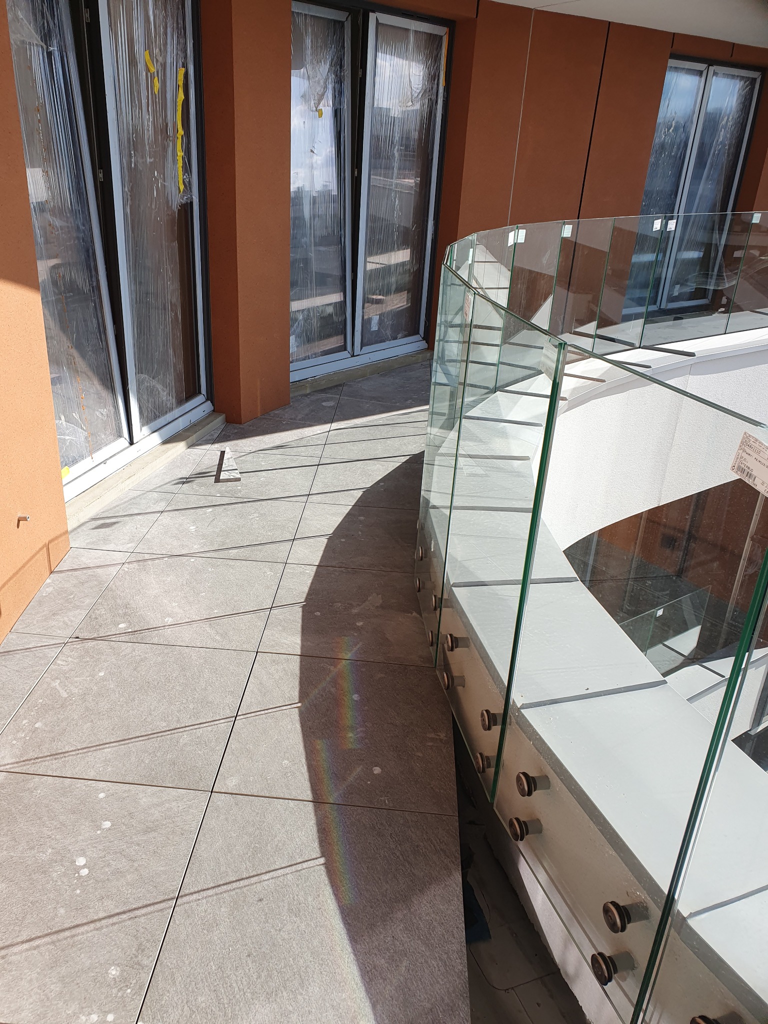 Wyłożony płytkami taras z szklaną balustradą, widoczne okna balkonowe w folii ochronnej i cień rzucany przez konstrukcję