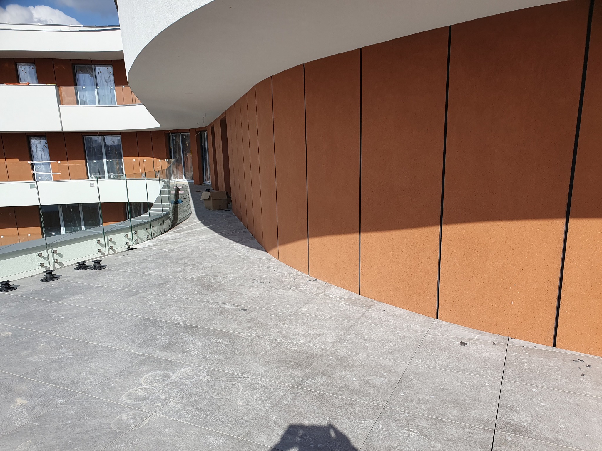 Wykończony taras z szarych płytek, brązowa elewacja budynku z widocznymi oknami i szklaną balustradą.