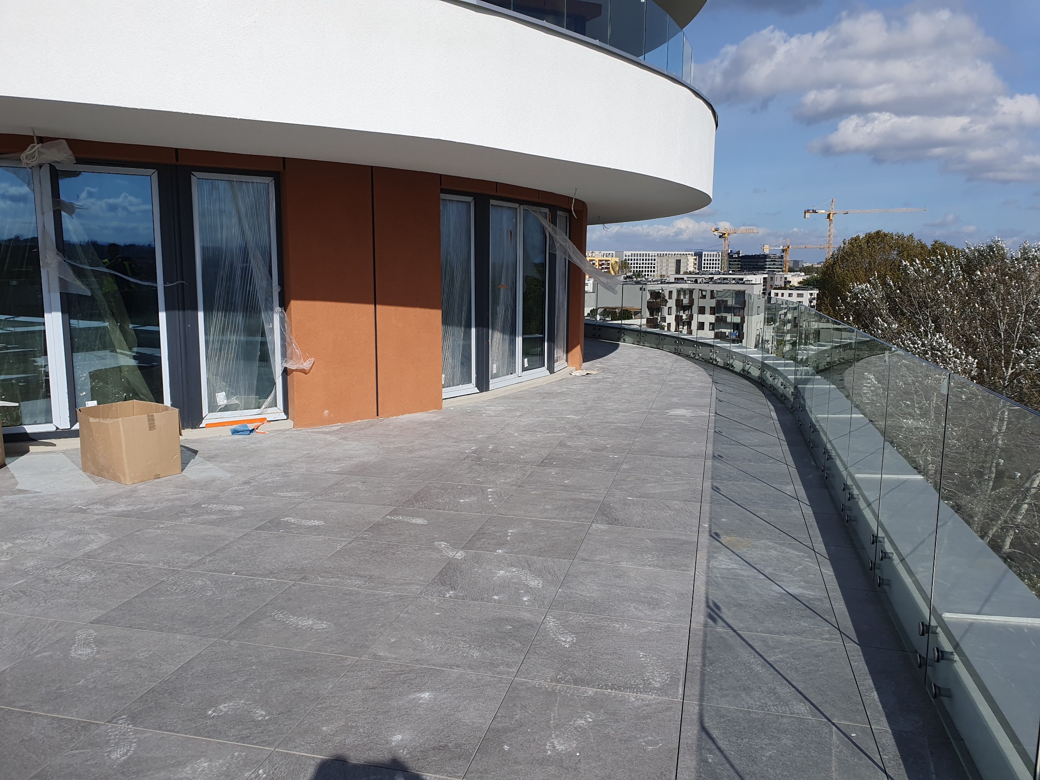 Wyłożony szarymi płytkami taras balkonu w nowoczesnym budynku z widokiem na miasto, szklane balustrady, okna zabezpieczone folią ochronną, widoczne ślady po pracach wykończeniowych.