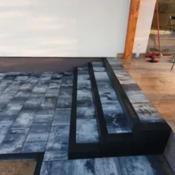 Niedokończone schody z kostki brukowej w odcieniach szarości, z czarnymi obrzeżami, prowadzące do tarasu; widoczny fragment elewacji budynku z oknem.