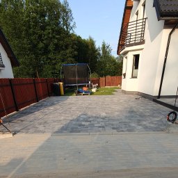 KomarBruk Łukasz Białobrzewski - Nowo ułożona, szara kostka brukowa na podjeździe przed domem z ogrodem. Widoczny fragment ogrodzenia i trampolina w tle. Jasne, naturalne światło.