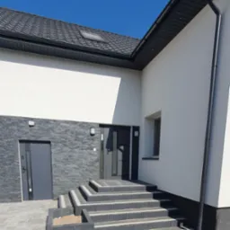 Elewacja budynku z kamienną okładziną, szare drzwi wejściowe, schody z kostki brukowej i donica. Błękitne niebo w tle. Nowoczesny design.