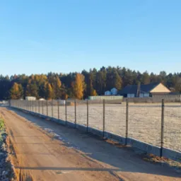 Długa, metalowa siatka ogrodzeniowa z betonową podmurówką, ciągnąca się wzdłuż polnej drogi w kierunku lasu i zabudowań w słoneczny poranek.