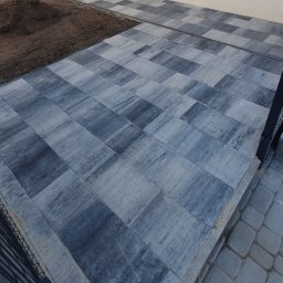 KomarBruk Łukasz Białobrzewski - Ułożona szara kostka brukowa w geometryczny wzór, przylegająca do ściany budynku i obszaru z ziemią. Fragment ogrodzenia w prawym dolnym rogu kadru.