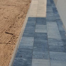 KomarBruk Łukasz Białobrzewski - Ścieżka z szarej kostki brukowej ułożona wzdłuż budynku, z widocznym gruntem i ogrodzeniem w tle. Ujęcie z perspektywy, podkreślające regularność wzoru.