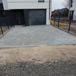 KomarBruk Łukasz Białobrzewski - Szara kostka brukowa na podjeździe nowoczesnego domu z garażem. Widoczne obrzeża, ogrodzenie panelowe i fragment instalacji drenażowej. Częściowo przygotowany teren pod dalsze prace.