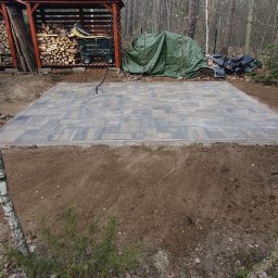 KomarBruk Łukasz Białobrzewski - Ułożona kostka brukowa na przygotowanym podłożu, widok z góry. W tle skład drewna i zieleń. Kostka w odcieniach szarości i brązu, ułożona w regularny wzór.