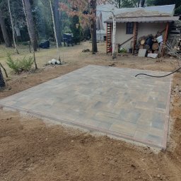 KomarBruk Łukasz Białobrzewski - Ułożona kostka brukowa na przygotowanym podłożu, obrzeża z czerwonej kostki. W tle drzewa i budynek gospodarczy.