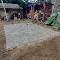 KomarBruk Łukasz Białobrzewski - Ułożona kostka brukowa w odcieniach szarości i brązu, obramowana czerwoną kostką. W tle drewniana wiata na drewno i budynek gospodarczy.