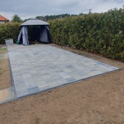 KomarBruk Łukasz Białobrzewski - Ukończony taras z kostki brukowej w odcieniach szarości, obrzeża z ciemnej kostki. W tle altana ogrodowa i żywopłot. Widok z góry, pochmurne niebo.