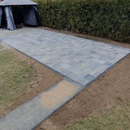 KomarBruk Łukasz Białobrzewski - Szara kostka brukowa ułożona na posesji, widoczny fragment ścieżki z kostki i altana ogrodowa w tle. Częściowo przygotowany teren pod dalsze prace.