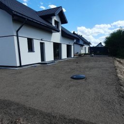 KomarBruk Łukasz Białobrzewski - Przygotowany teren pod budowę z widocznymi domami w tle, wyrównana ziemia, studzienka kanalizacyjna. Niebo z chmurami.
