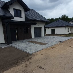 KomarBruk Łukasz Białobrzewski - Szara kostka brukowa ułożona przed nowoczesnym domem z ciemną stolarką i dachem. Fragment rabaty z kamykami. Widoczne ślady prac ziemnych.