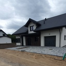 KomarBruk Łukasz Białobrzewski - Nowoczesny dom z szarą kostką brukową na podjeździe i trawnikiem w tle. Widoczne detale wykończenia budynku i otoczenia, w tym rury drenażowe.