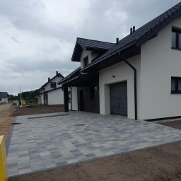 KomarBruk Łukasz Białobrzewski - Ułożona szara kostka brukowa na podjeździe do nowoczesnego domu z garażem. Widoczne obrzeża i przygotowany teren pod dalsze prace.