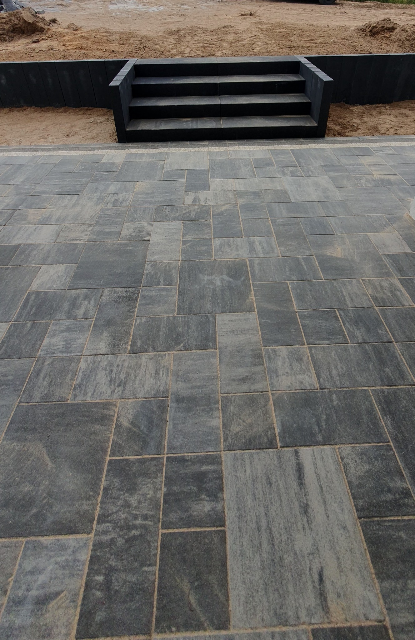 Ułożona szara kostka brukowa w geometryczny wzór, prowadząca do schodów z ciemnych płyt na tle piaszczystego terenu. Widok z dołu.