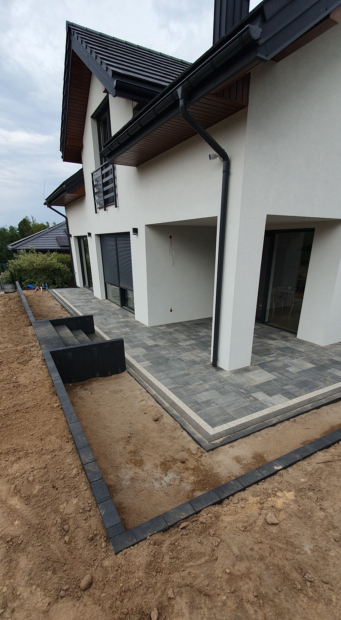 Nowoczesny taras z kostki brukowej w odcieniach szarości, z jasną obwódką. Widoczne schody i fragment ogrodu w trakcie realizacji.