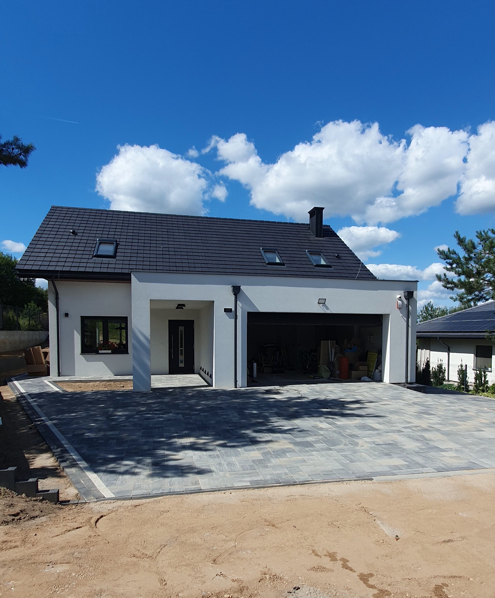 Nowoczesny dom z podjazdem z szarej kostki brukowej ułożonej we wzór. Widoczne fragmenty niezakończonych prac ziemnych. Błękitne niebo z białymi chmurami w tle.
