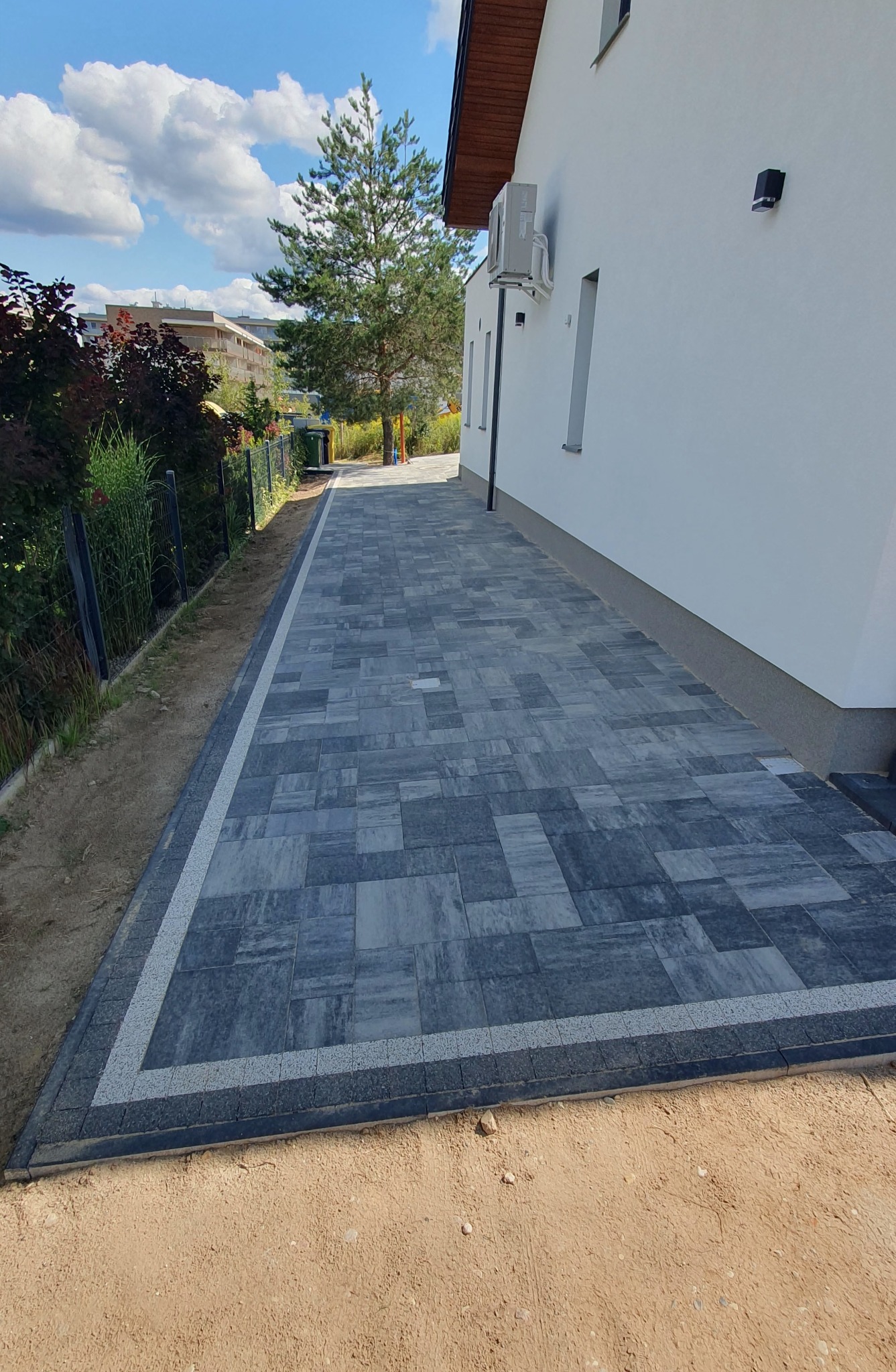 Elegancka ścieżka z szarej kostki brukowej, obramowana jasnym pasem, prowadząca wzdłuż białej ściany domu z widocznym klimatyzatorem i czujnikiem ruchu.