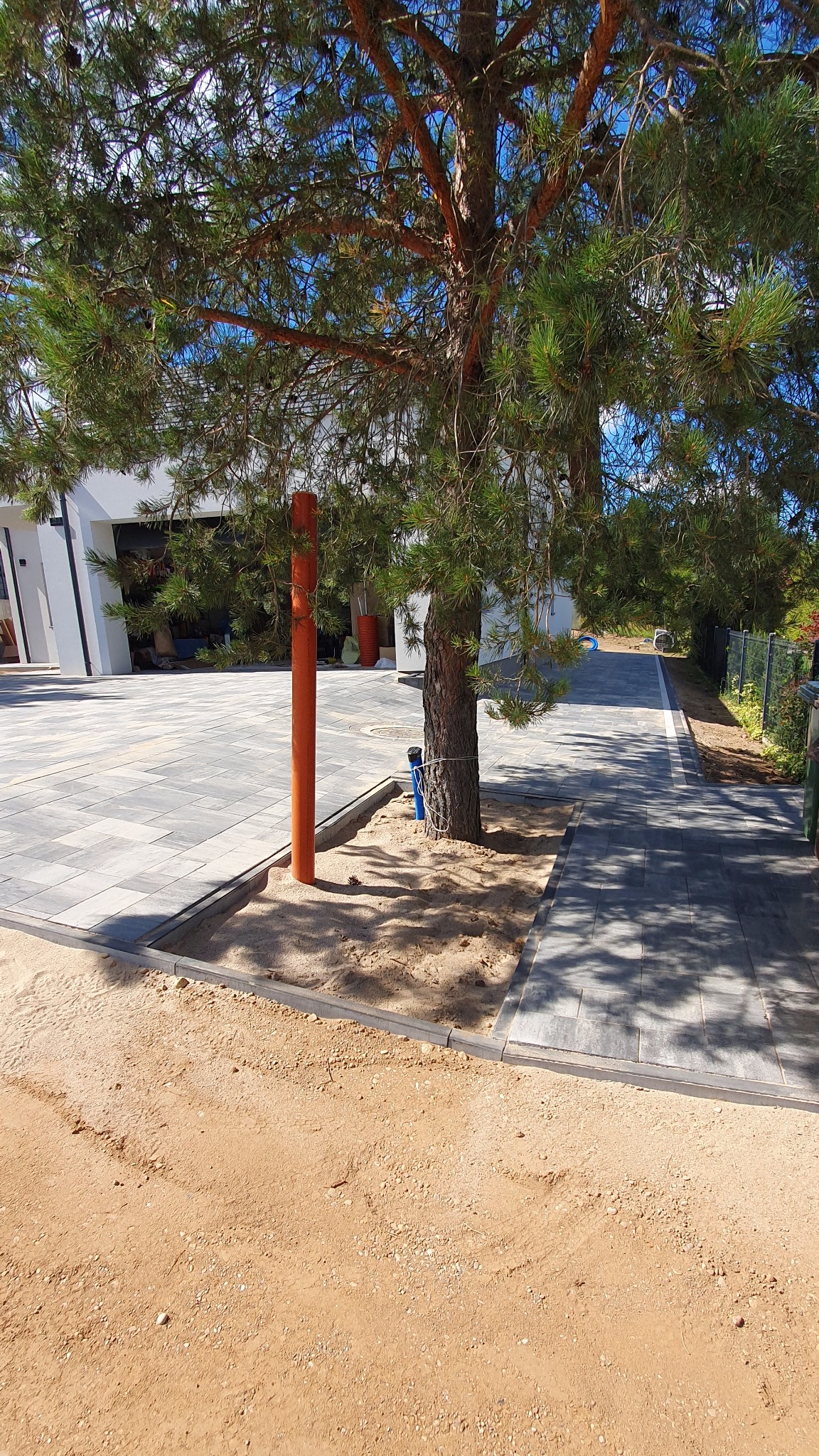 Nowoczesna posesja z szarej kostki brukowej, obrzeża, drzewo w kwadracie z piasku, pomarańczowa rura. Jasne, słoneczne oświetlenie.