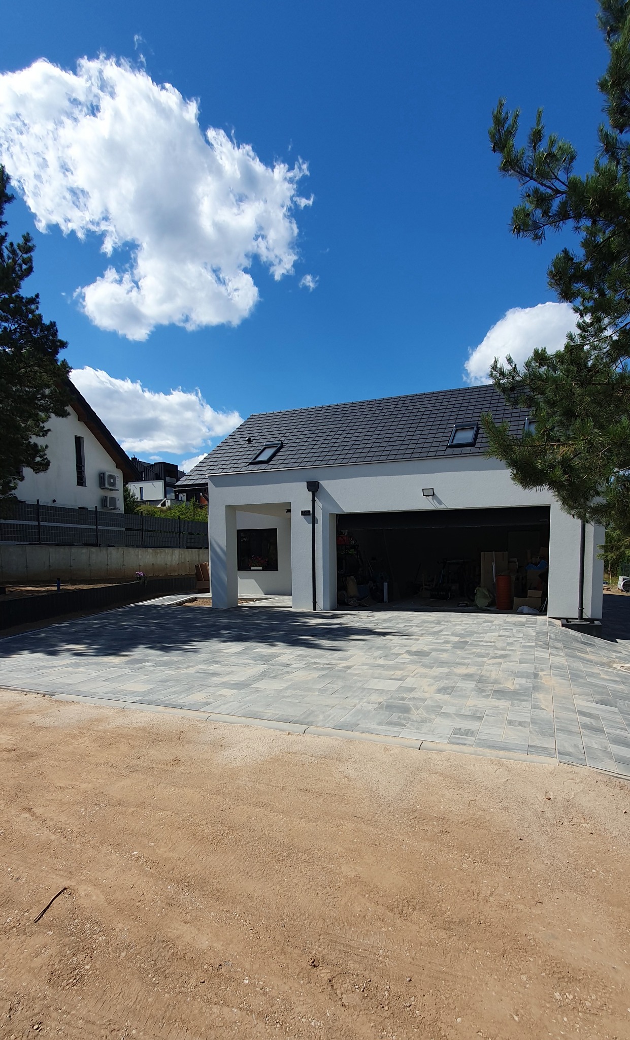 Nowoczesny garaż z ułożoną, szarą kostką brukową na podjeździe. Widoczne jasne niebo z chmurami i fragmenty zieleni. Kostka ułożona w geometryczny wzór.