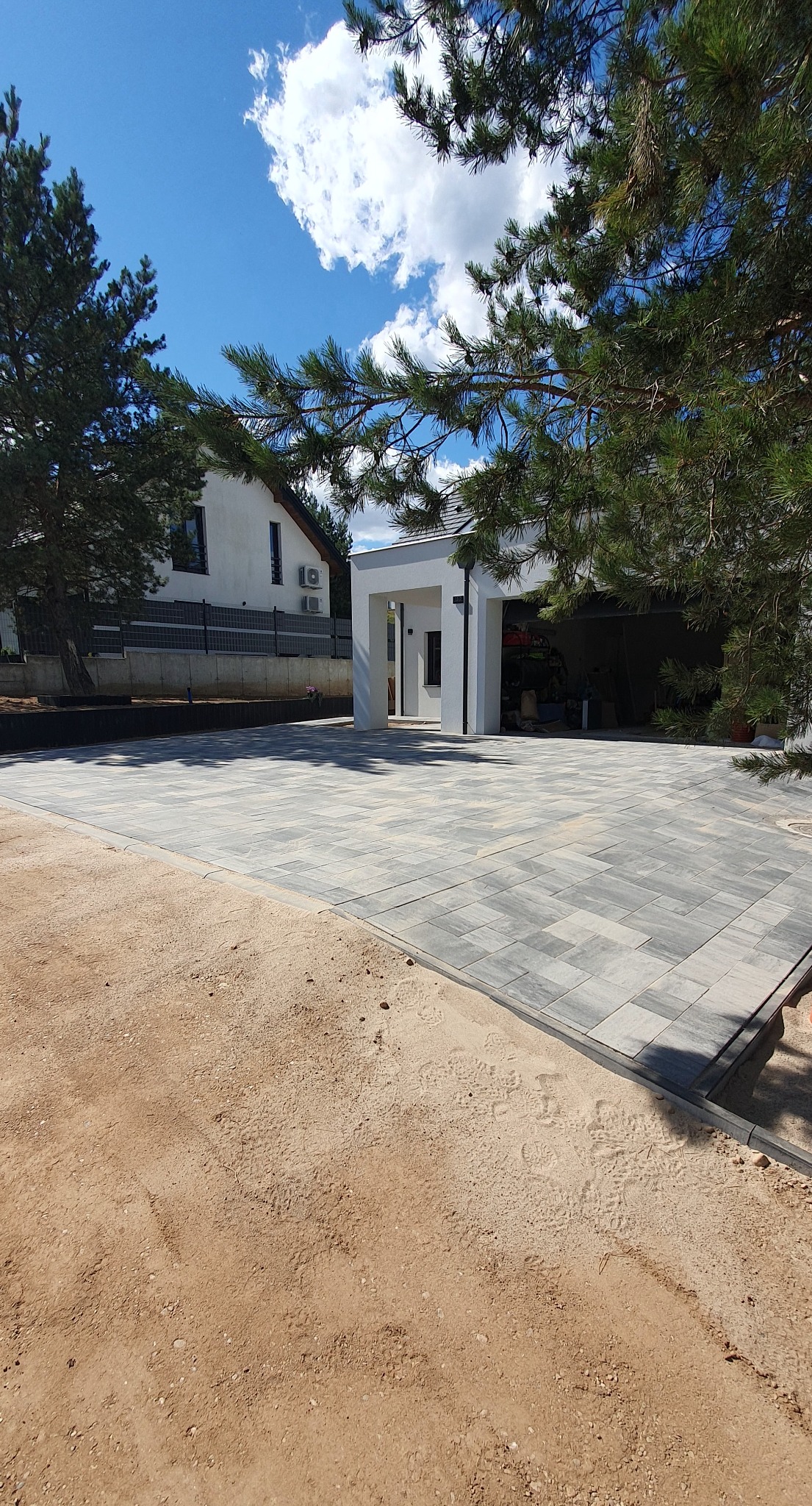 Ułożona szara kostka brukowa na posesji domu z białym garażem, widok z poziomu gruntu, fragment piasku i gałęzi drzewa w kadrze, błękitne niebo z chmurami.