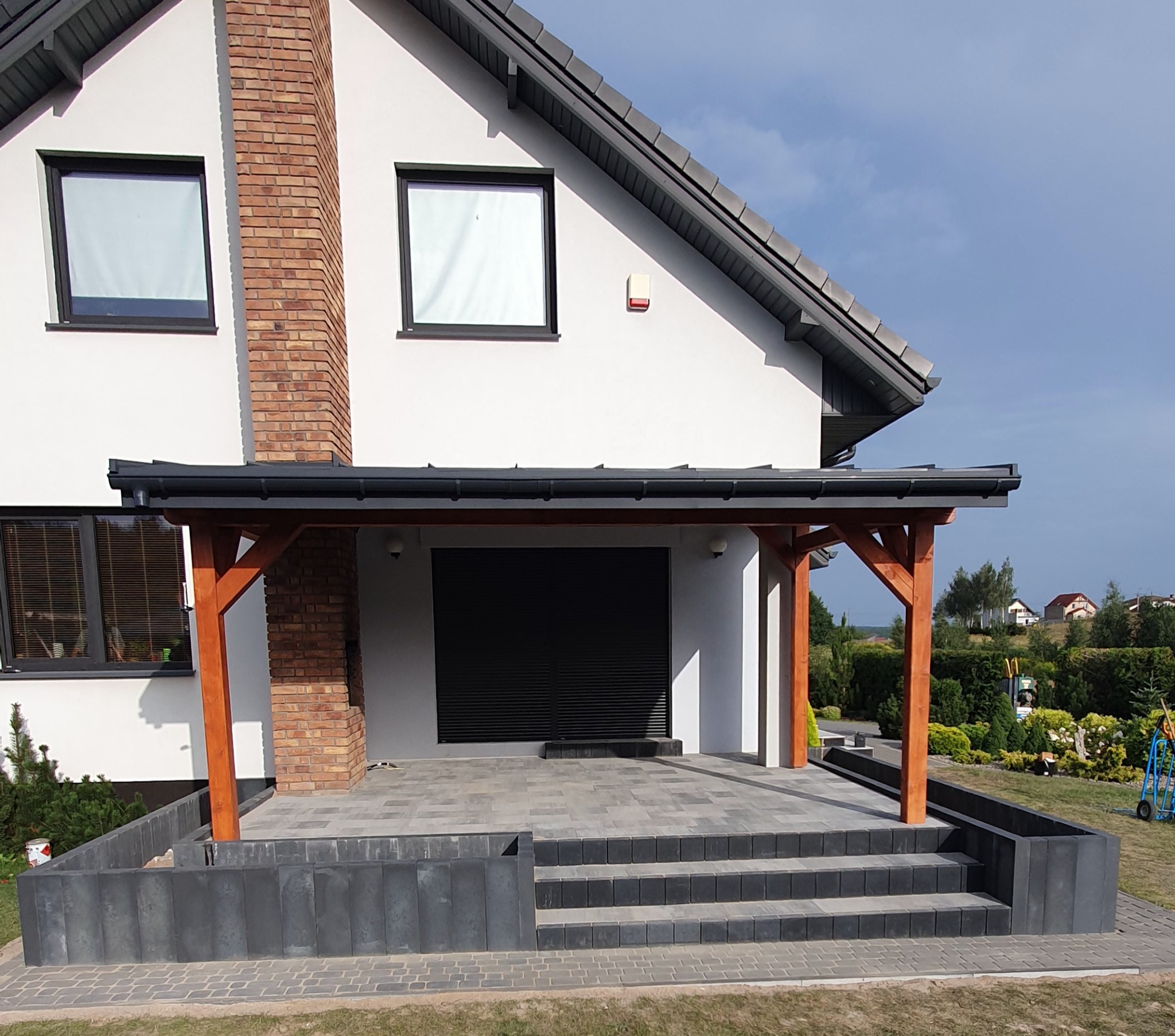 Nowoczesny taras z kostki brukowej przy domu z drewnianą konstrukcją zadaszenia i schodami z ciemnoszarej kostki. Widok na ogród i okoliczne zabudowania.