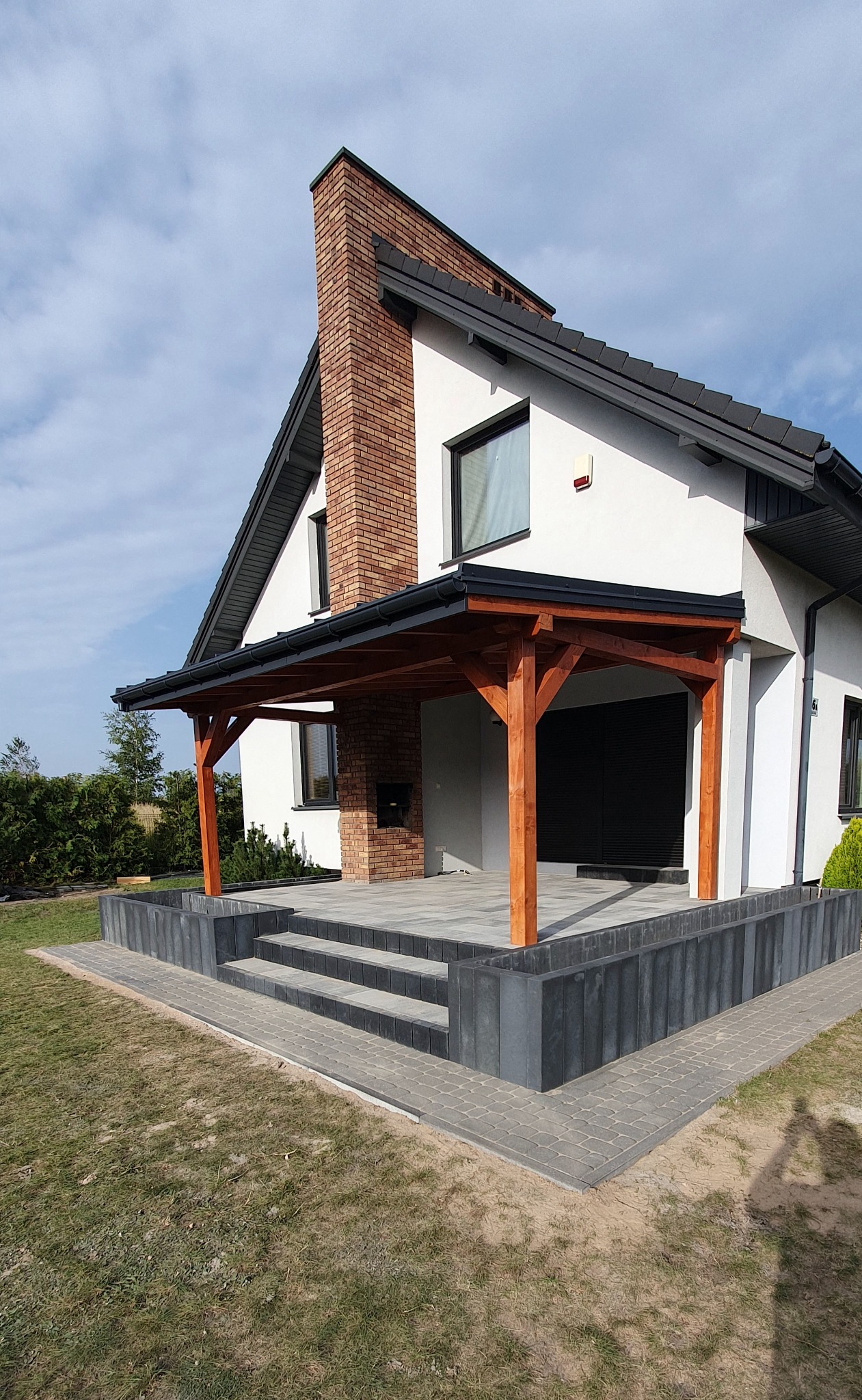 Nowoczesny dom z tarasem z kostki brukowej i drewnianą pergolą, widok z zewnątrz. Schody z kostki prowadzą na taras. Kompozycja z ceglanym kominem.