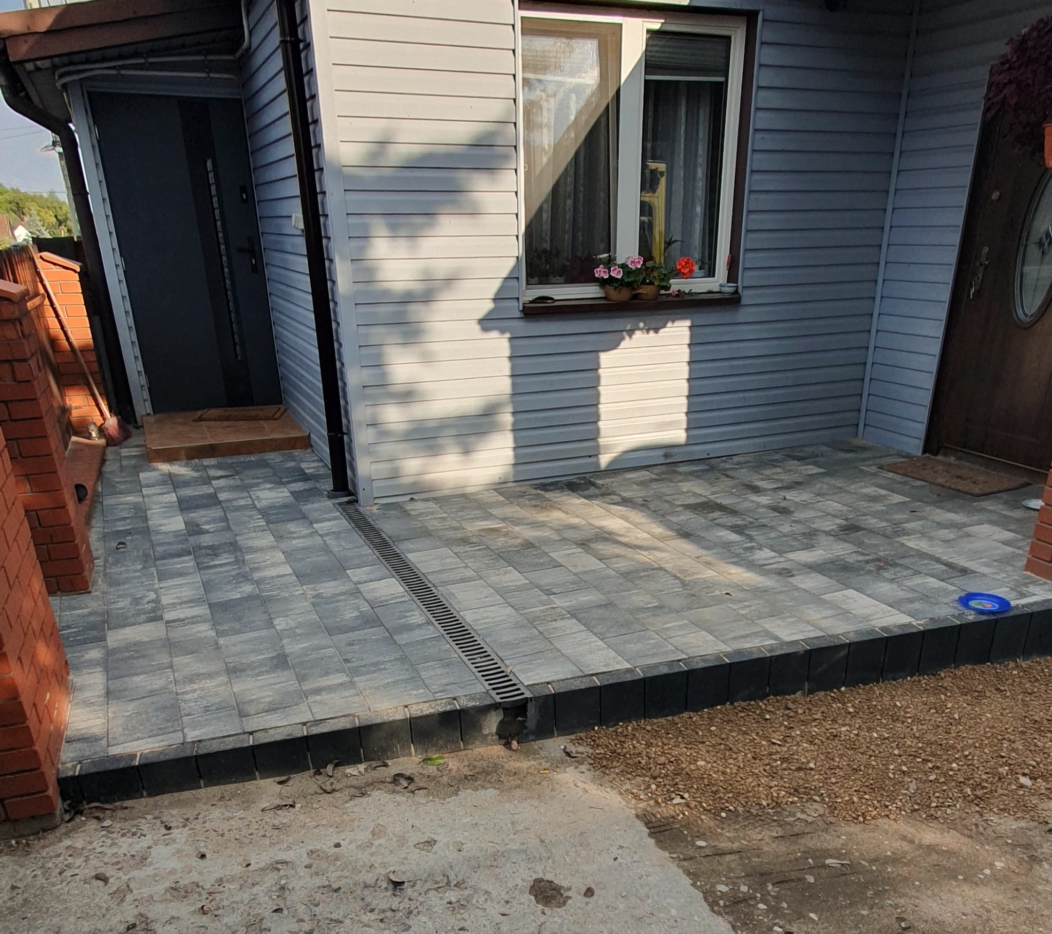 Szara kostka brukowa ułożona przed domem z szarą elewacją, widoczny drenaż liniowy i fragment ceglanego murku. Ujęcie z niskiego kąta, światło słoneczne.