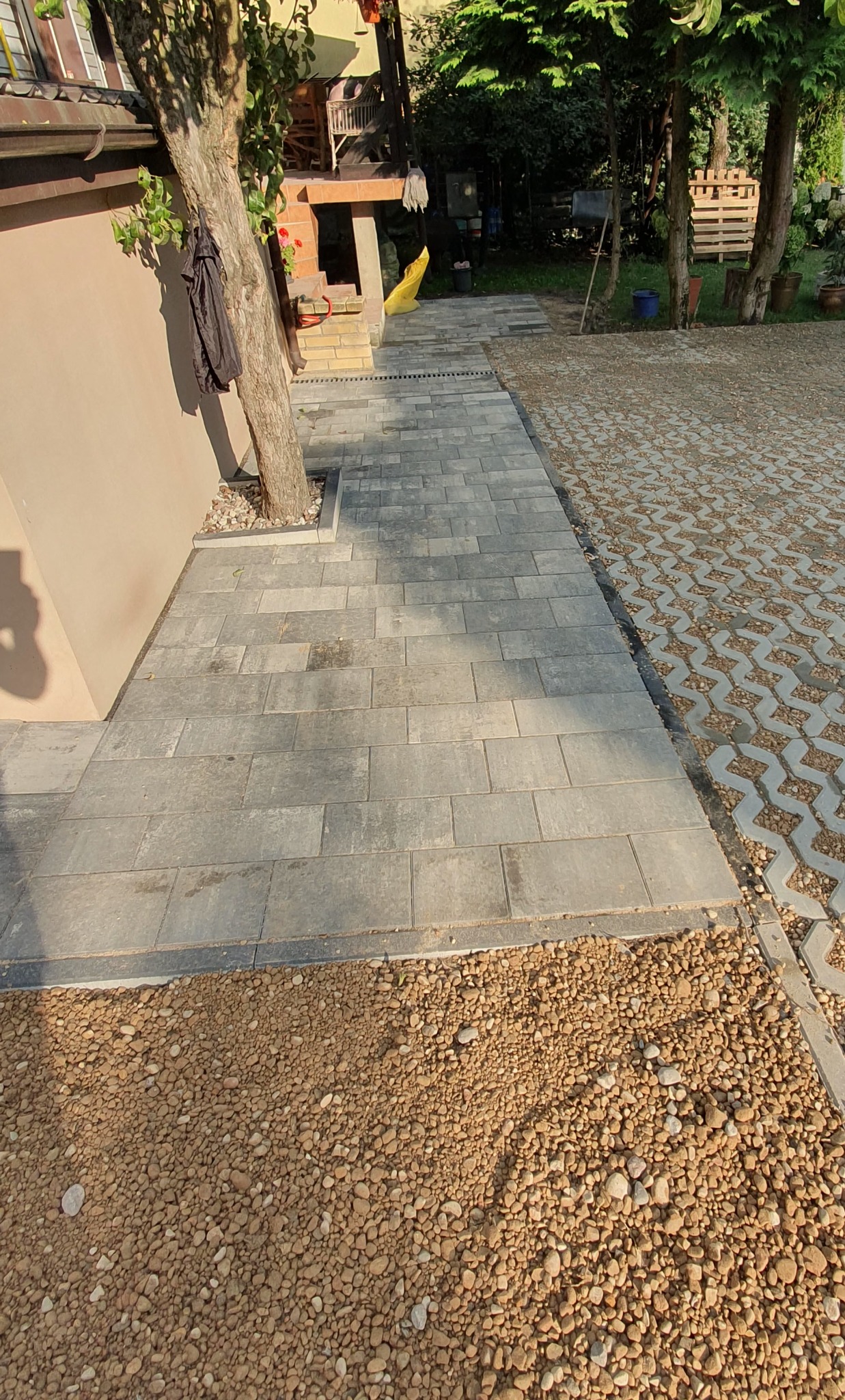 Ułożona ścieżka z kostki brukowej obok budynku, przechodząca w ażurowy parking. Wykończenie z drobnych kamieni. Widok na zieleń ogrodu.