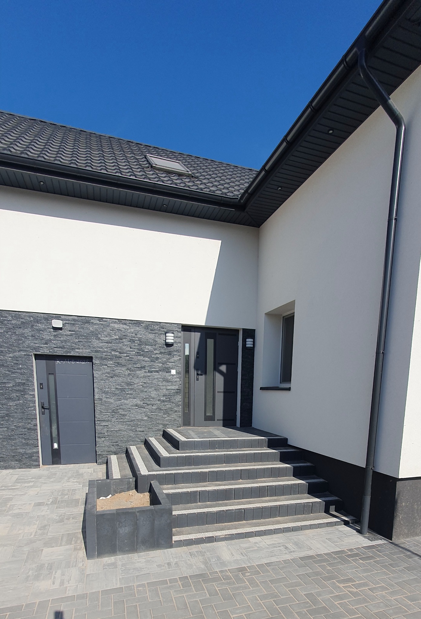 Elewacja budynku z kamienną okładziną, szare drzwi wejściowe, schody z kostki brukowej i donica. Błękitne niebo w tle. Nowoczesny design.