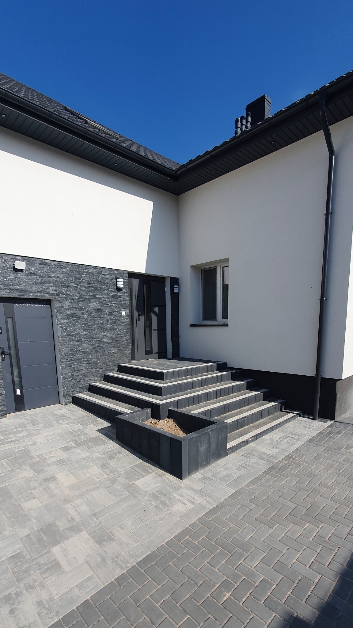 Nowoczesne schody wejściowe z kostki brukowej w odcieniach szarości, prowadzące do domu z białymi ścianami i elementami z kamienia. Widoczna rabata z kostki brukowej.
