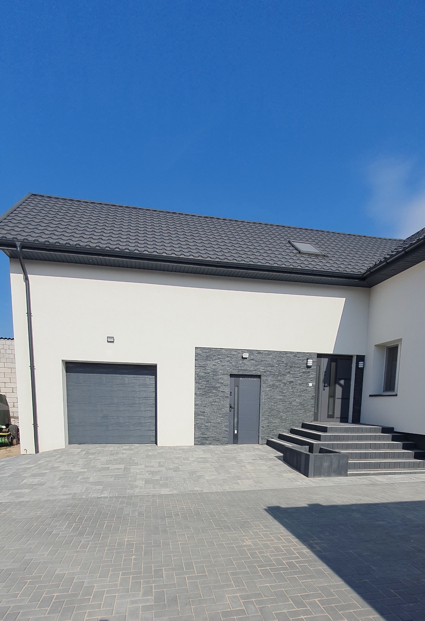 Nowoczesny dom z szarą kostką brukową ułożoną w jodełkę. Wejście z kamiennymi schodami i szarą stolarką. Błękitne niebo w tle.