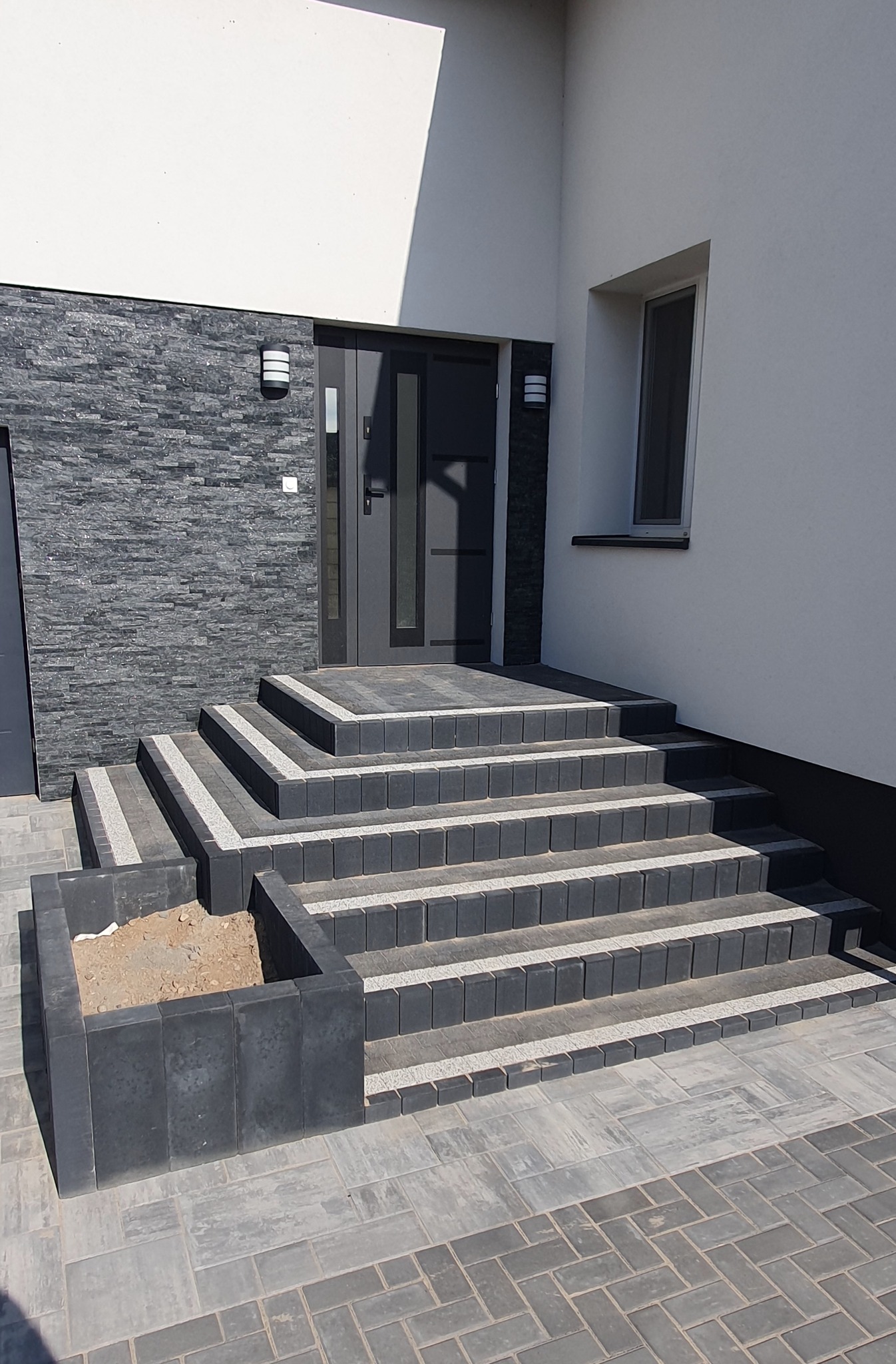 Nowoczesne schody wejściowe z ciemnej kostki brukowej z jasnymi akcentami, prowadzące do drzwi. Obok schodów donica z kostki. Całość na tle jasnej elewacji.