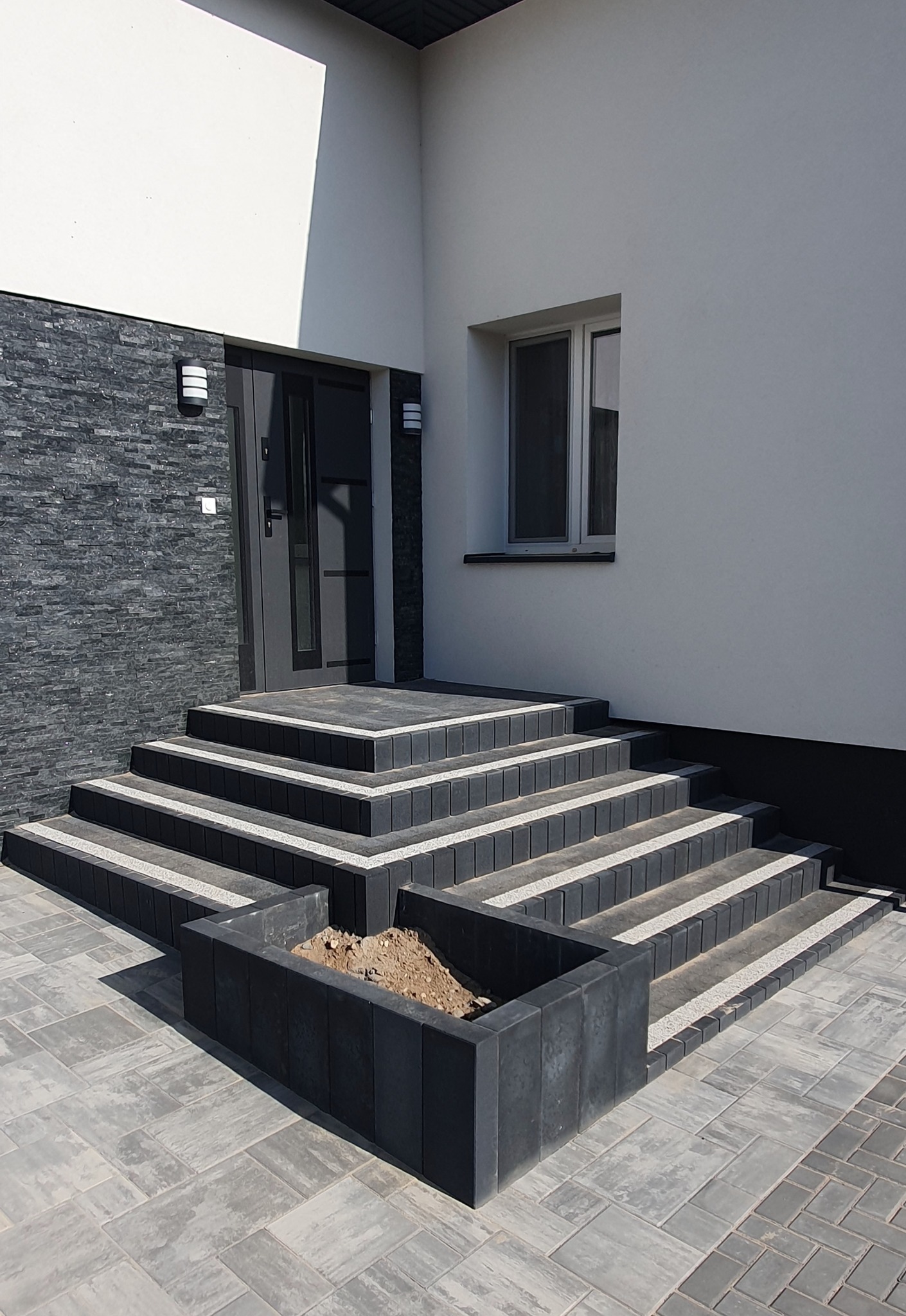Nowoczesne schody wejściowe z kostki brukowej w odcieniach szarości, z elementami dekoracyjnymi i donicą. Minimalistyczna elewacja budynku w tle.