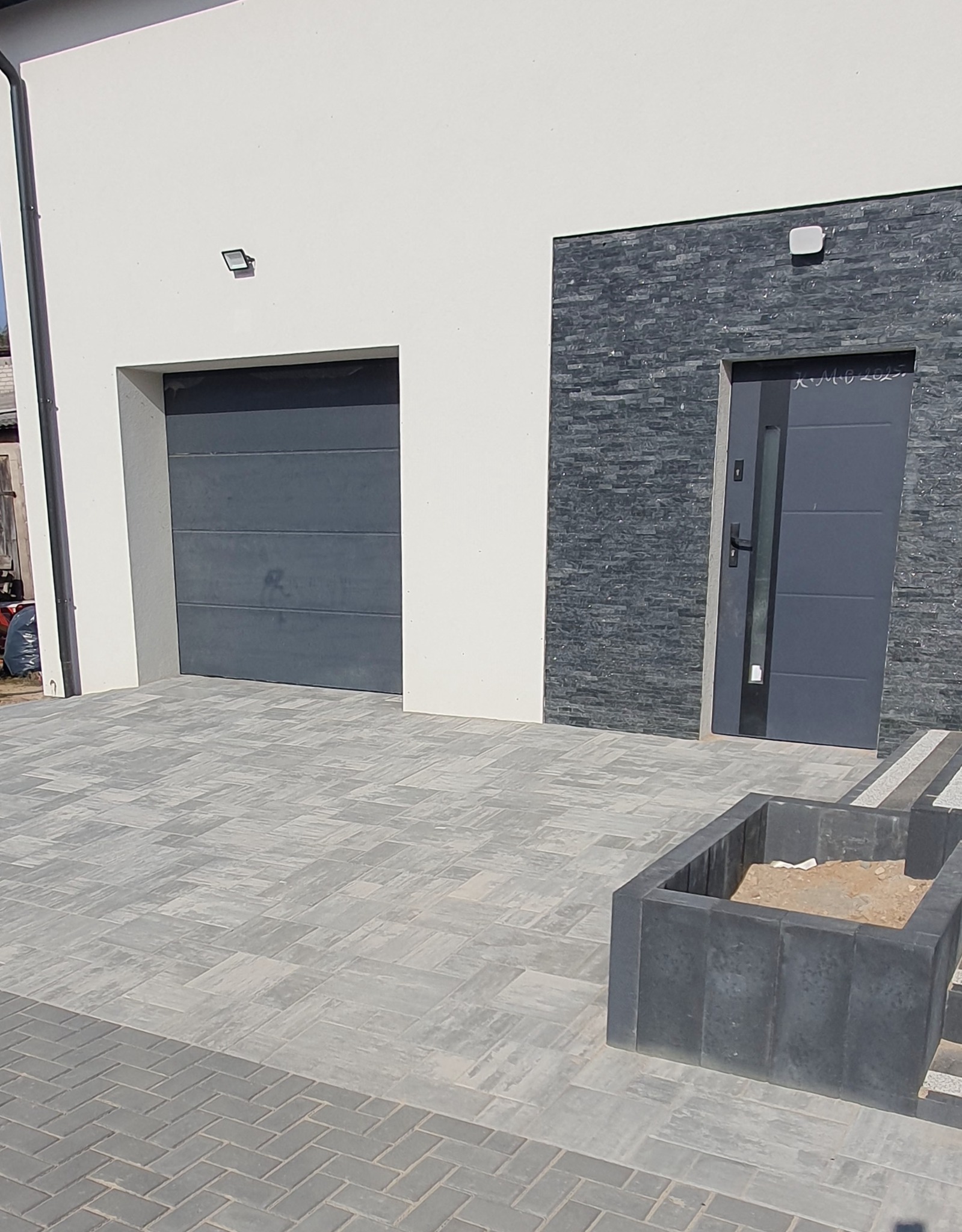 Ułożona kostka brukowa w szarych odcieniach przed budynkiem z garażem i wejściem wykończonym kamieniem. Widoczna donica z piaskiem.
