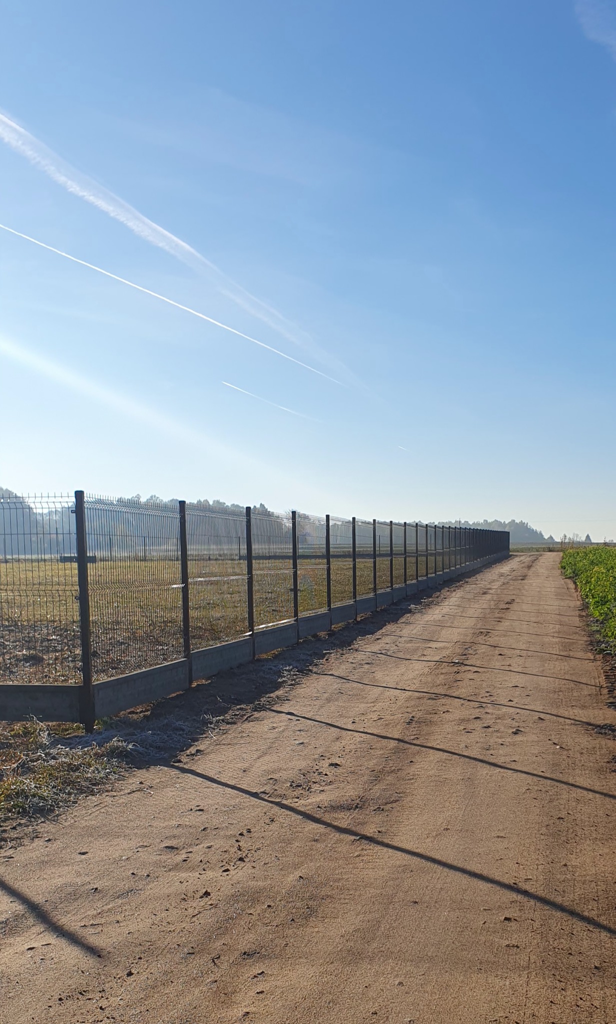 Długa, metalowa siatka ogrodzeniowa z betonową podmurówką, ciągnąca się wzdłuż polnej drogi w słoneczny dzień. Widoczne ślady smug kondensacyjnych na błękitnym niebie.