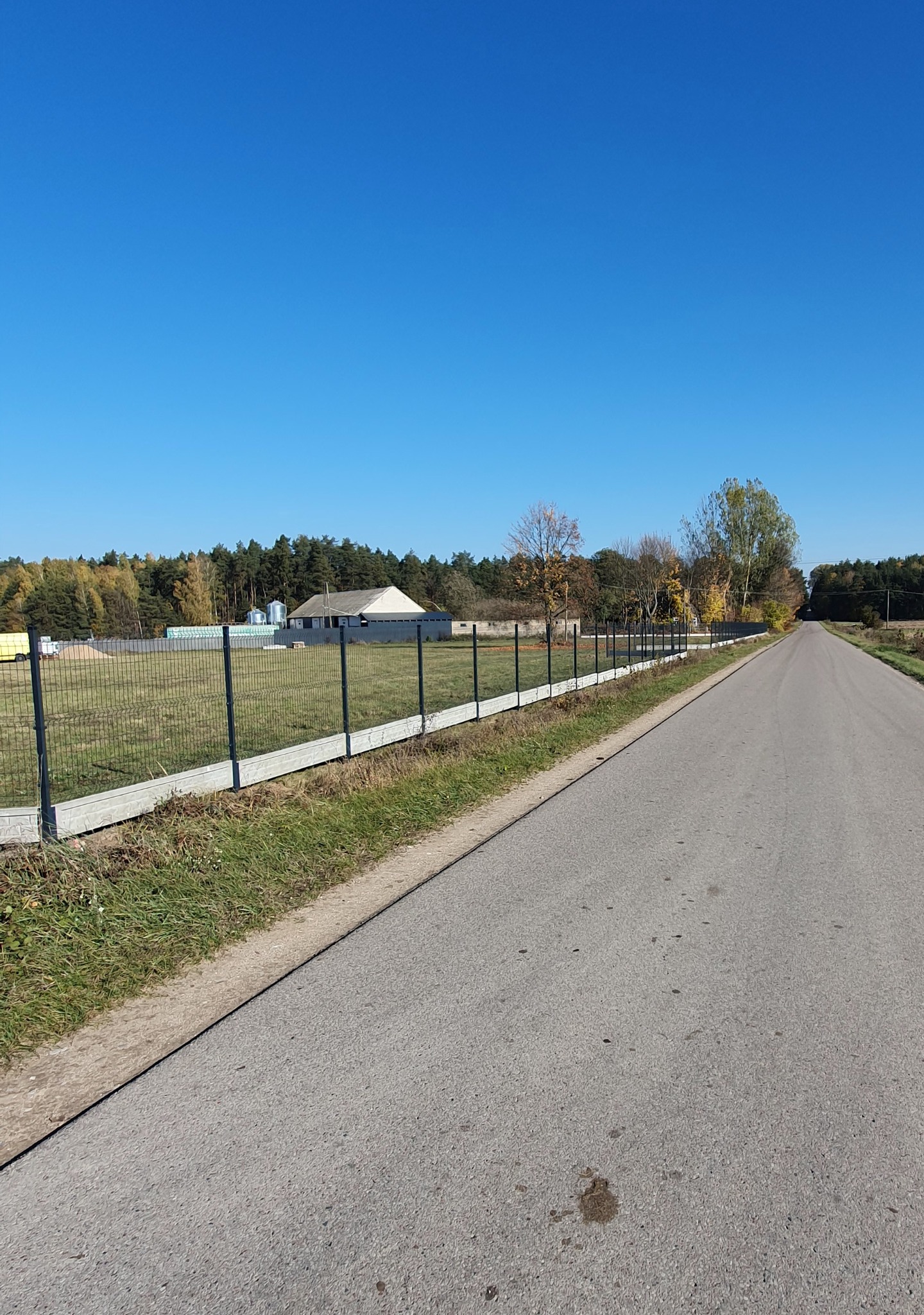 Długa, prosta droga asfaltowa obok ogrodzenia panelowego z podmurówką betonową, w tle budynki gospodarcze i las, słoneczny dzień.
