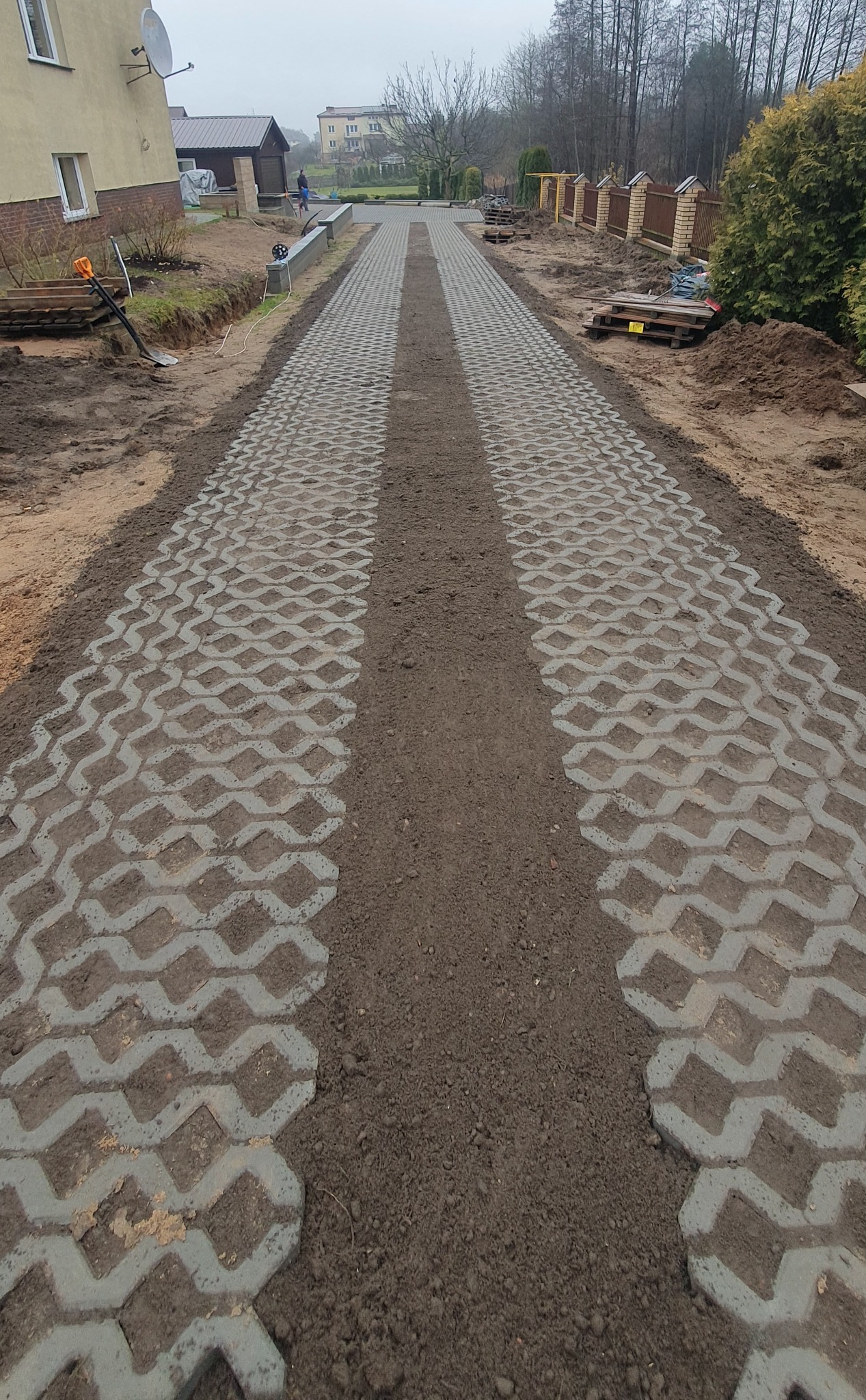 Długa, częściowo ułożona droga z ażurowych, szarych płyt betonowych. W tle widoczny budynek i fragment ogrodzenia. Prace brukarskie w toku.