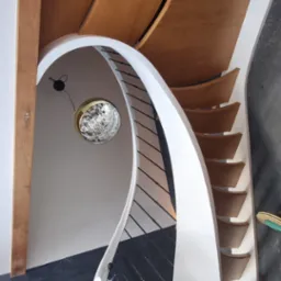 Spiralne schody z drewnianymi stopniami i białymi policzkami, czarne metalowe balustrady, widok z góry, ozdobna lampa wisząca.