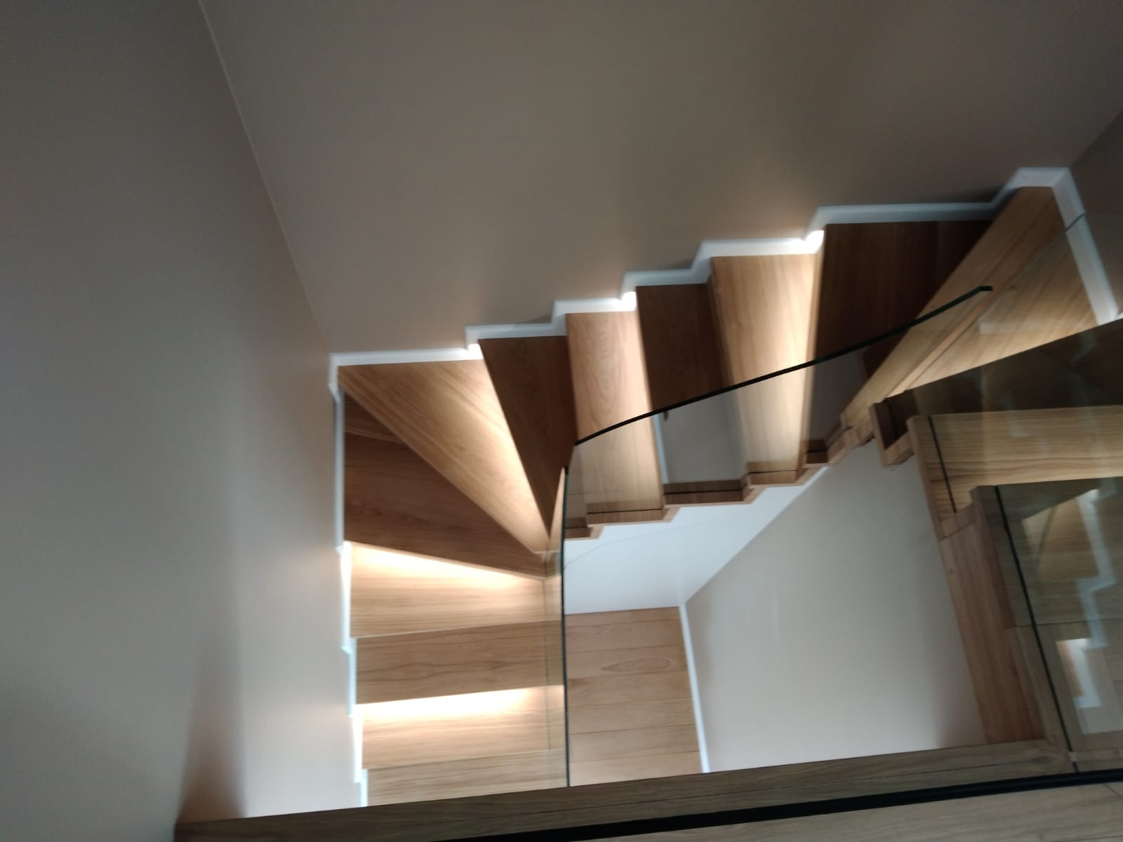 Kątowe ujęcie schodów z drewna z podświetleniem LED, zabezpieczonych szklaną balustradą, widok z góry.