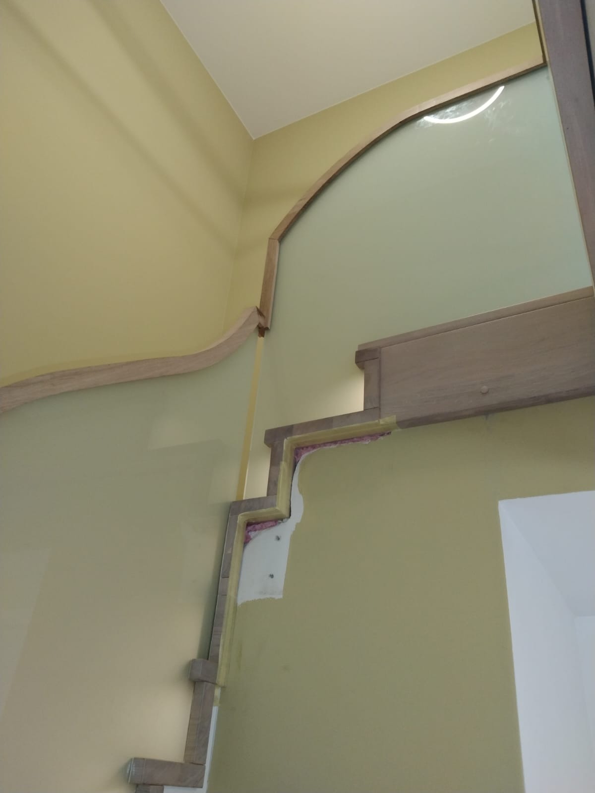 Wnętrze w trakcie prac wykończeniowych, widoczne fragmenty konstrukcji schodów z drewnianymi elementami i oświetleniem, ściany w odcieniach żółci i zieleni, widoczne materiały izolacyjne.