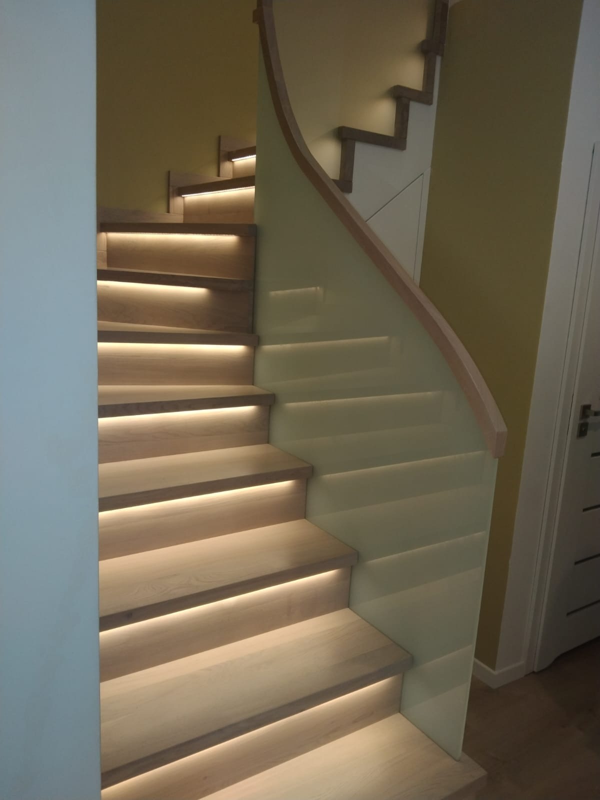 Nowoczesne schody z jasnego drewna z podświetleniem LED pod każdym stopniem i szklaną balustradą z drewnianym poręczą.
