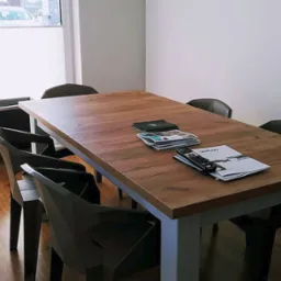 Wnętrze biura z drewnianym stołem konferencyjnym, krzesłami o geometrycznym designie oraz ulotkami i pilotem na blacie. W tle widoczne okno z żaluzjami plisowanymi i logo firmy Haiku Studio.