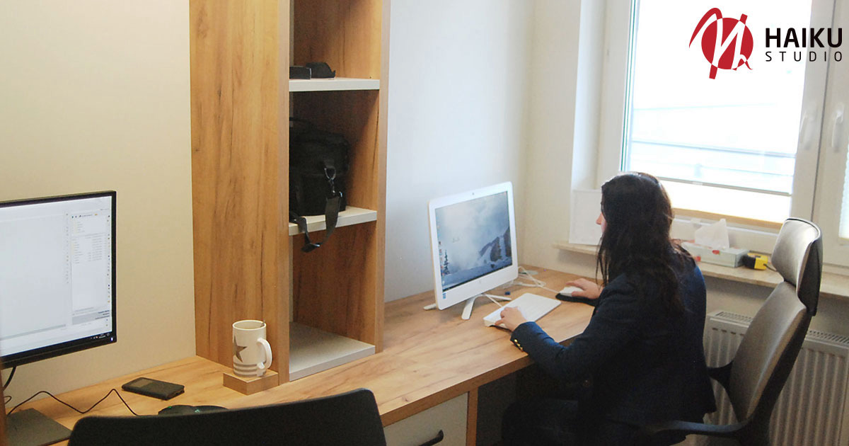 Kobieta w biurze pracuje przy biurku z dwoma monitorami, na jednym z nich widoczny kod źródłowy, obok kubek z gwiazdą, na półce torba i inne akcesoria biurowe, w tle okno z widocznym logo firmy.