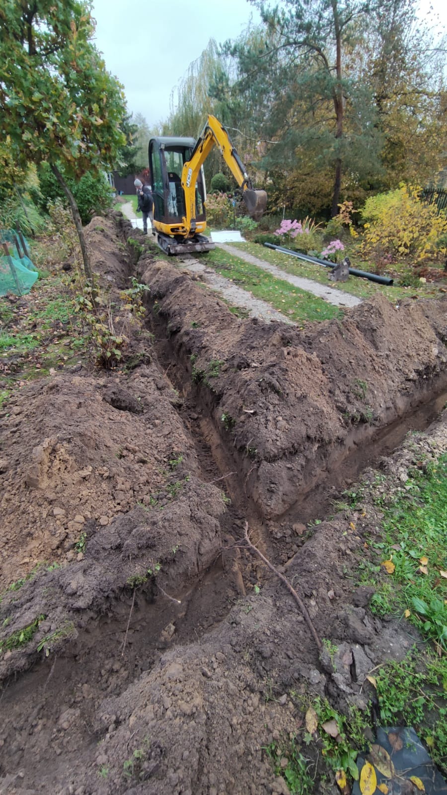 Żółta mini koparka JCB na trawniku obok wykopanego rowu o głębokich ścianach, w tle widoczna osoba i czarne rury.