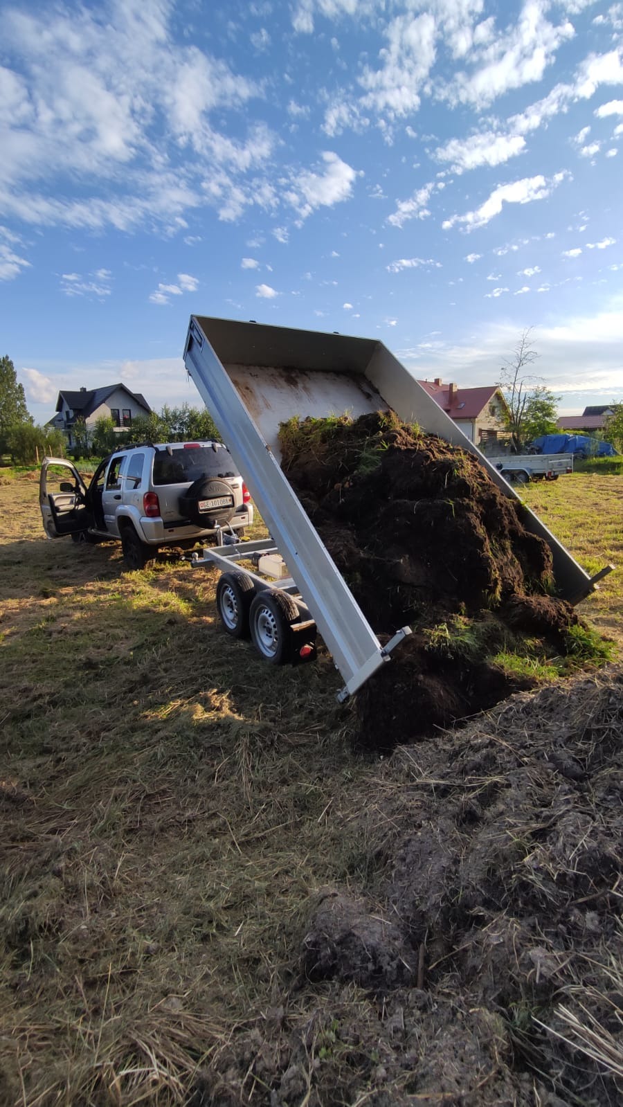 Przyczepa wywrotka z podniesioną skrzynią, wysypująca ziemię z trawą na pole; w tle częściowo widoczny samochód terenowy z otwartymi drzwiami oraz zabudowania.