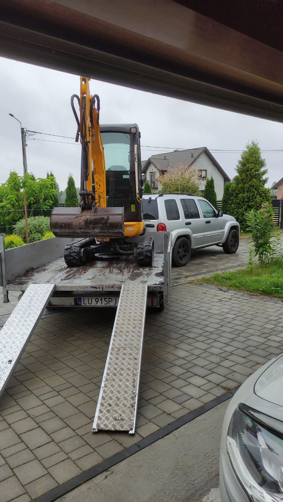 Żółta mini koparka na platformie przyczepy, z rampami najazdowymi, zaparkowana na brukowanym podjeździe przed domem i srebrnym SUVem.
