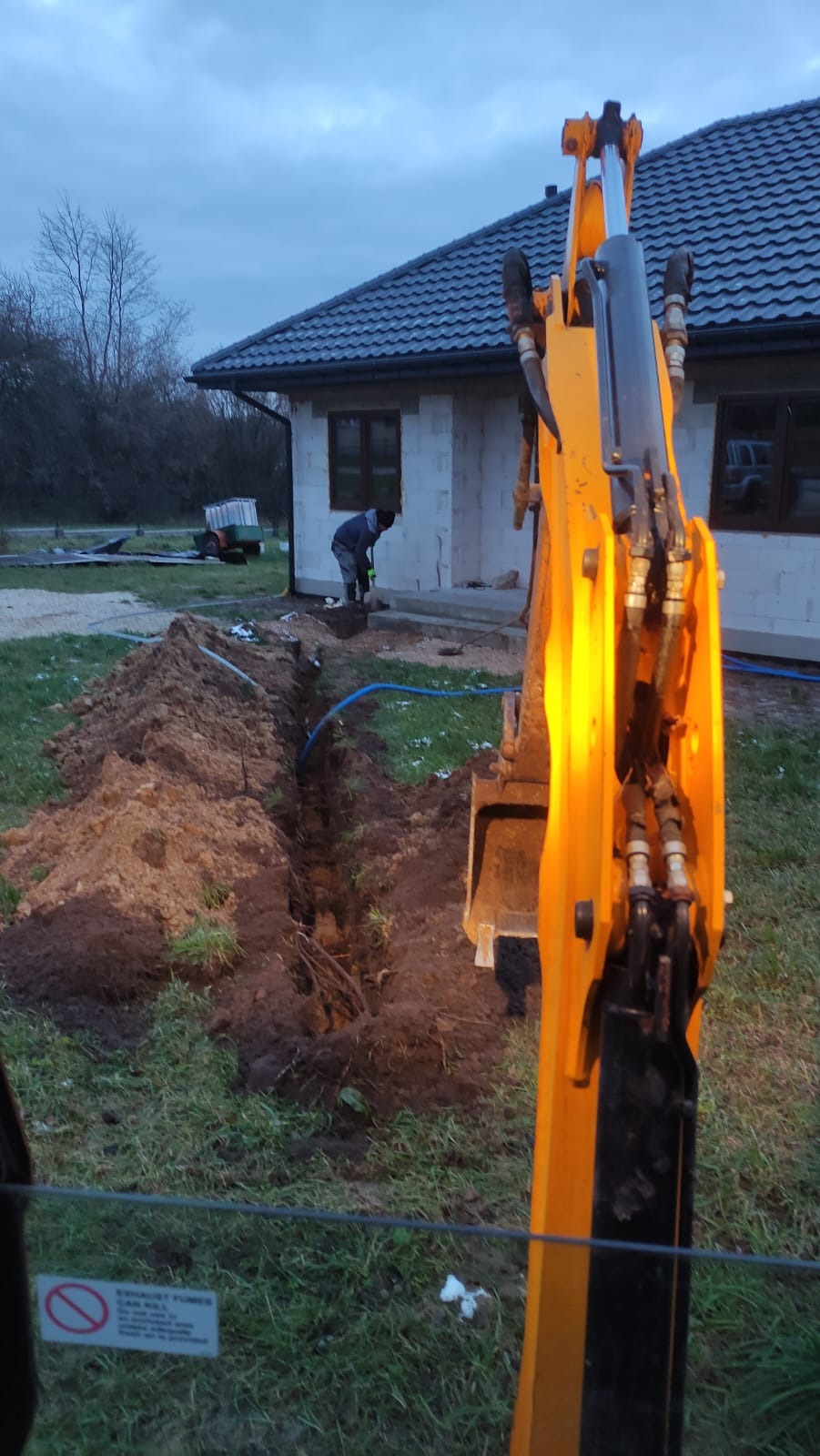 Żółta koparka na pierwszym planie, wykopany wąski rów z niebieską rurą w ziemi, pracownik z łopatą przy fundamentach domu w tle, pochmurne niebo.